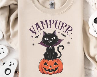 May include: Beige sweatshirt with a Halloween-themed graphic. The design features a black cat wearing a purple cape, sitting atop an orange jack-o'-lantern. The word "VAMPURR" is arched above the cat in purple lettering, with bats and stars.
