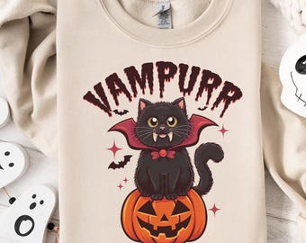 May include: A beige sweatshirt featuring a Halloween-themed graphic. The design includes a black cat dressed as a vampire, sitting on an orange jack-o'-lantern. The word "VAMPURR" is arched above the cat in a dark red font.