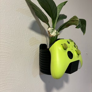 Könnte beinhalten: Ein leuchtend grüner Videospiel-Controller ist an einer Wand mit einem schwarzen Halter befestigt. Hinter dem Controller befindet sich eine Topfpflanze mit grünen Blättern.