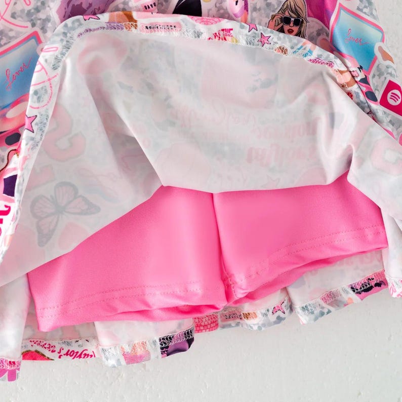 May include: A skort featuring a white outer layer with a colorful collage print of butterflies, hearts, stars, and illustrated figures, paired with a solid bright pink inner short layer. The fabric has a smooth texture and a visible hemline.