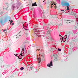 May include: A fabric featuring a collage of pink and white graphics including hearts, butterflies, smiley faces, guitars, disco balls, and text such as "it's me, hi! I'm the problem, it's me," "Lover," "1989," "Taylor's version," and "meet me in the afterglow."