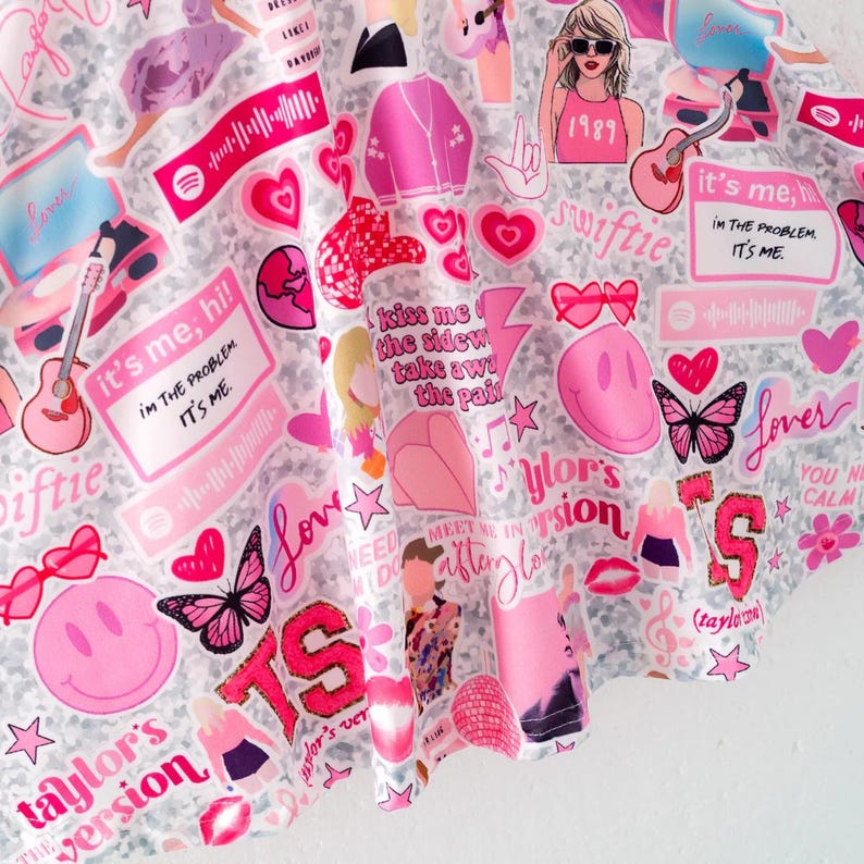 May include: A fabric featuring a collage of pink and white graphics including hearts, butterflies, smiley faces, guitars, disco balls, and text such as "it's me, hi! I'm the problem, it's me," "Lover," "1989," "Taylor's version," and "meet me in the afterglow."