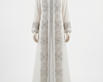 Op de afbeelding: Een vloerlange, gebroken witte kaftan met lange mouwen en een open voorzijde. Het kledingstuk is versierd met ingewikkeld zilverkleurig borduurwerk en heldere kristallen die verticaal langs de voorpanden en op de manchetten zijn aangebracht.