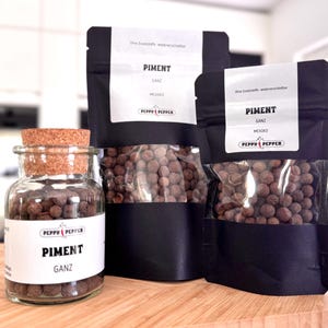 Pode incluir: Três recipientes de bagas de pimenta da Jamaica inteiras. Os recipientes são pretos com janelas de plástico transparente e etiquetas brancas que dizem "Piment Ganz" e "Peppo Pepper".