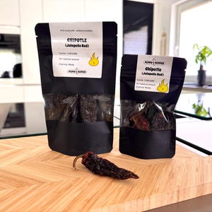 Può includere: Due buste nere con pannelli anteriori trasparenti contenenti peperoncini jalapeño rossi secchi etichettati "Chipotle (Jalapeno Red)" con un punteggio di piccantezza di 5. Le buste sono su una superficie di legno con un singolo peperoncino jalapeño rosso secco.