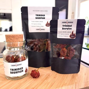 Op de afbeelding: Drie zakjes gedroogde Trinidad Scorpion pepers, een zeer hete chilipeper. De zakjes zijn zwart met witte tekst en een rood vlamlogo. De Scoville-schaal is 500.000 tot 1.380.000 SHU. Een zakje is open en laat de gedroogde pepers zien. Een klein glazen potje met een kurken deksel bevat ook gedroogde pepers. Een enkele rode peper ligt op het houten oppervlak.