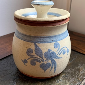 May include: A ceramic jar with a lid, featuring a cream-colored base with blue and brown accents. The jar is adorned with a blue floral and bird design. The lid has a small handle. The jar is sitting on a dark surface.