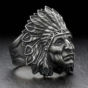 Puede incluir: Un anillo plateado que presenta una detallada representación de un jefe nativo americano con un tocado de plumas. El anillo tiene intrincadas tallas y se encuentra sobre un fondo oscuro, destacando la detallada artesanía de la joyería.