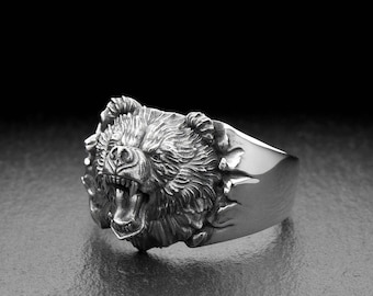 May include: A silver-colored ring featuring a detailed bear head with an open mouth and visible teeth. The bear's fur is textured, and the ring has a wide band. The ring is set against a dark background.