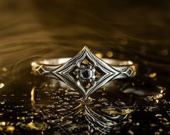 May include: A silver ring with a geometric design and a blue gemstone in the centre. The ring is sitting on a black surface with water droplets.