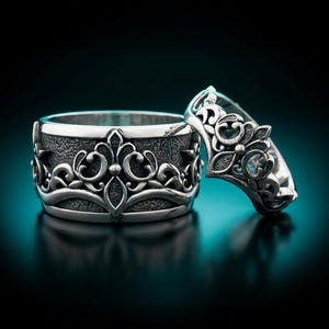 Puede incluir: Dos anillos de plata con diseños florales ornamentados. Uno es una banda ancha con un patrón detallado, el otro un anillo más pequeño con un diseño similar y una pequeña piedra preciosa. Los anillos se presentan sobre un fondo oscuro.