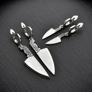 May include: Silver-toned knife earrings and matching smaller knife earrings. The earrings feature a detailed knife blade and handle design. The jewelry is displayed on a dark, textured surface, creating a striking contrast.