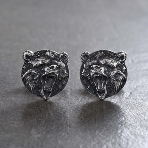 以下が含まれることがあります： 銀色のクマの頭のカフスボタン2つ。各カフスボタンは、歯を見せる開いた口を持つ詳細なクマの頭が特徴です。クマの頭は丸く、質感のある金属仕上げです。背景は灰色の表面です。