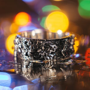 May include: A silver-colored cuff bracelet with a detailed floral and leaf pattern. The bracelet is in sharp focus, with a blurred background of colorful bokeh lights. The design features holly leaves and berries, suggesting a holiday theme.
