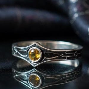 A cinematic close-up of a sterling silver ring with a distinct "V" shaped or "Lance Tip" contour. The focal point is a bezel-set, round gemstone with a deep amber-orange hue (Citrine or Madeira Citrine). The shoulders of the band feature horizontal grooves that taper toward the center, resembling the shaft of a spear or the fletching of an arrow. The metal is polished but features dark oxidation in the grooves for contrast.