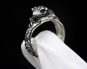 Pode incluir: Um anel de metal prateado com uma figura de sapo esculpida que repousa sobre o topo da banda. A banda tem uma aparência texturizada semelhante a um ramo, com detalhes oxidados escuros nas fendas, criando um acabamento de alto contraste.