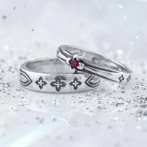 May include: Two silver rings with intricate floral designs. One ring features a small, red gemstone. The rings are displayed on a white surface with scattered glitter, creating a sparkling effect. Ideal for weddings or engagements.