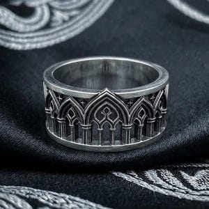 May include: A silver ring with an intricate gothic arch design. The ring is set against a dark, patterned fabric, highlighting its detailed architecture. The ring's design features repeating arches and vertical lines.