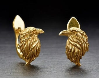 May include: A pair of gold-colored earrings, each featuring a detailed raven head design. The earrings have a post and clip closure. The raven's feathers are intricately carved, and the earrings are set against a dark background.
