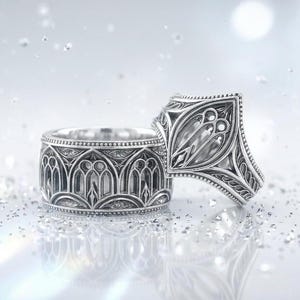 May include: Two silver rings with intricate gothic arch designs. One ring is upright, showcasing the detailed pattern, while the other lies on its side. The rings are set against a bright, blurred background with sparkling accents.