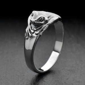 May include: A silver ring with a frog design. The frog is sculpted on the ring's top, with visible eyes and limbs. The ring is set against a dark background, emphasising its metallic shine. The ring is likely made of sterling silver.