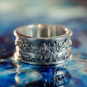Può includere: Un anello d'argento con una rappresentazione dettagliata dell'Ultima Cena. L'anello è su uno sfondo blu e bianco.