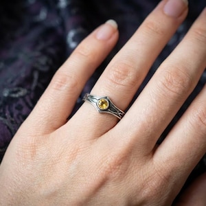 The ring is bathed in a warm, golden mist, creating a magical, ethereal atmosphere. The reflection on the wet surface below mirrors the amber glow of the stone. The mood is heroic and legendary, evoking the feeling of a magical artifact discovered in a dragon's lair or an elven armory. It appeals to fans of Lord of the Rings or high-fantasy RPGs.