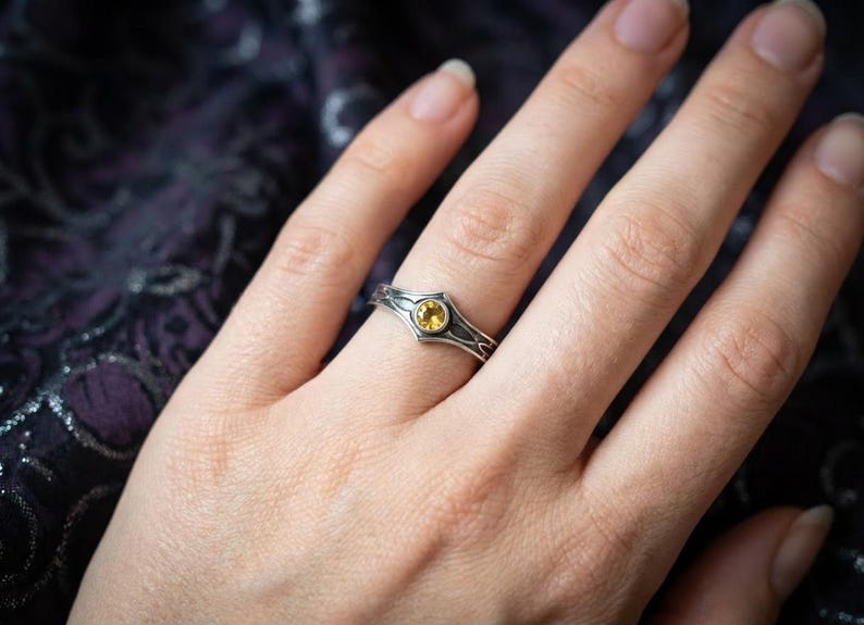 The ring is bathed in a warm, golden mist, creating a magical, ethereal atmosphere. The reflection on the wet surface below mirrors the amber glow of the stone. The mood is heroic and legendary, evoking the feeling of a magical artifact discovered in a dragon's lair or an elven armory. It appeals to fans of Lord of the Rings or high-fantasy RPGs.