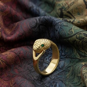 Pode incluir: Anel dourado em forma de cobra, com cabe&ccedil;a texturizada e uma pequena gema verde. Descansa sobre um tecido estampado em tons de bord&ocirc;, verde e ouro. O design evoca uma serpente.