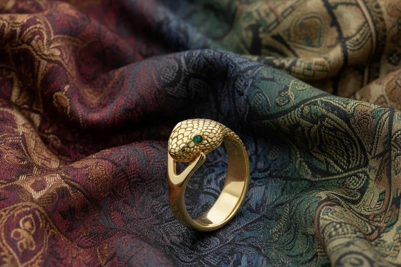 Pode incluir: Anel dourado em forma de cobra, com cabe&ccedil;a texturizada e uma pequena gema verde. Descansa sobre um tecido estampado em tons de bord&ocirc;, verde e ouro. O design evoca uma serpente.