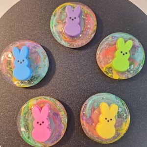 以下が含まれることがあります： イースターバニーの樹脂カボション5個。各カボションには、紫、青、緑、ピンク、黄色の異なる色のウサギがいます。ウサギは、渦巻くカラフルな樹脂の背景に配置されています。