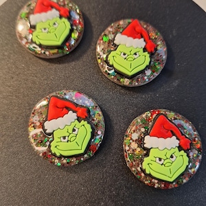 May include: Four round, glittery Christmas-themed embellishments featuring the Grinch in a Santa hat. The Grinch is green with a red and white hat. The glitter is a mix of colours, including red, green, silver, and gold.