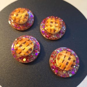 May include: Four round resin pieces with miniature pie designs. Each pie has a lattice crust and sits atop a bed of glitter in shades of red, pink, and gold. The resin is clear, allowing the glitter to sparkle.