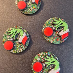 May include: Four round, Christmas-themed decorative buttons. Each button features a green hand with a red and white outfit, a red circle, and a glittery green background. The buttons are arranged on a dark grey surface.
