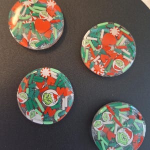 May include: Four round, clear resin Christmas-themed magnets. Each magnet features a festive design with green, red, and white sprinkles, Grinch faces, and heart shapes. The magnets are set against a dark background.