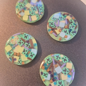 Könnte beinhalten: Vier runde, grüne Resin-Stücke mit Glitzereffekt. Jedes Stück ist mit kleinen, quadratischen und rechteckigen Formen in Grün, Braun, Gelb und Blau verziert. Das Design zeigt pixelige Gesichter.