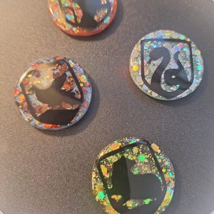May include: Four circular resin pins with glitter and black silhouettes. The pins feature a shield design with a lion, a snake, a badger, and a raven. The glitter is in various colours, including gold, green, and orange.