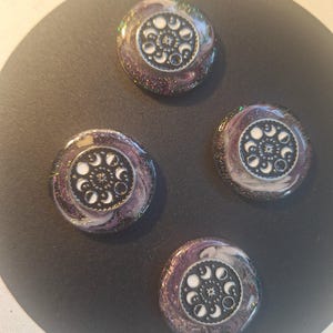 Puede incluir: Cuatro botones circulares con una incrustación central negra y gráficos de fases lunares en blanco. Cada botón está rodeado por un borde de resina brillante y arremolinada en tonos morado oscuro, negro y blanco con partículas de purpurina.