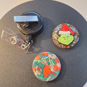 May include: A black retractable badge reel with a clear plastic strap, beside two round Christmas-themed badge reel designs. One features the Grinch in a Santa hat, and the other has a festive pattern with red and green elements.