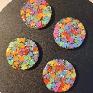 Puede incluir: Cuatro cabujones redondos de resina llenos de confeti en forma de corazón colorido. Los corazones son en tonos naranja, amarillo, verde, azul y morado, con acentos de purpurina. Los cabujones están sobre un fondo oscuro.
