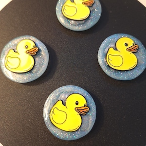 Puede incluir: Cuatro cabujones circulares de resina, cada uno con un gráfico de patito de goma amarillo brillante sobre un fondo azul claro translúcido incrustado con finas partículas de purpurina brillante en toda la base.