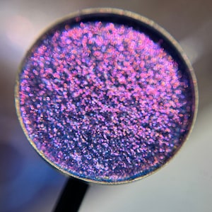 May include: Close-up of a round, metallic eyeshadow pan filled with shimmering, iridescent purple and pink pigments. The eyeshadow has a smooth, textured surface and is surrounded by a silver metal rim. A black handle is visible.