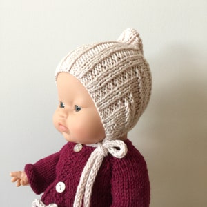 May include: A doll wearing a knitted beige bonnet with a tie. The doll is wearing a maroon knitted sweater.