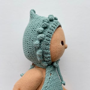 Può includere: Una bambola fatta a mano che indossa un berretto di lana blu chiaro con una punta a punta e una fila di pon pon attorno al bordo. Il berretto è legato sotto il mento con una sciarpa abbinata.