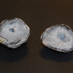 Op de afbeelding: Twee helften van een agaatgeode met een wit interieur en een donkergrijze buitenkant. De geodehelften liggen op een donkergrijs oppervlak.