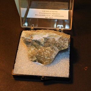 May include: A green and white gehlenite mineral specimen in a clear plastic display case. The label reads "Gehlenite, Crestmore Quarry, Riverside Co., California, M 183".