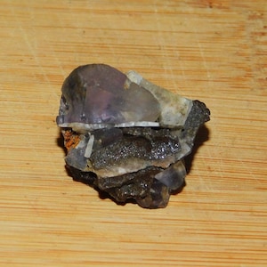May include: A purple and gray crystal with a layered structure, set against a wooden background.