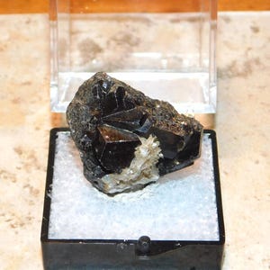 Puede incluir: Una taza de cerámica blanca con un asa negra y el texto "But First, Coffee" en negrita negra. La taza se encuentra sobre un fondo borroso, resaltando su diseño simple.