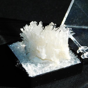 May include: A cluster of white crystal formations, resembling a burst of needles, sits within a black display case. The crystals are illuminated, showcasing their intricate structure. The case is partially open, revealing a clear, geometric structure.