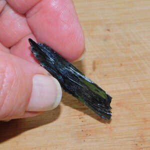 May include: A dark green and black crystal with a rough, jagged texture. The crystal is about 2 inches long and is held in a hand.